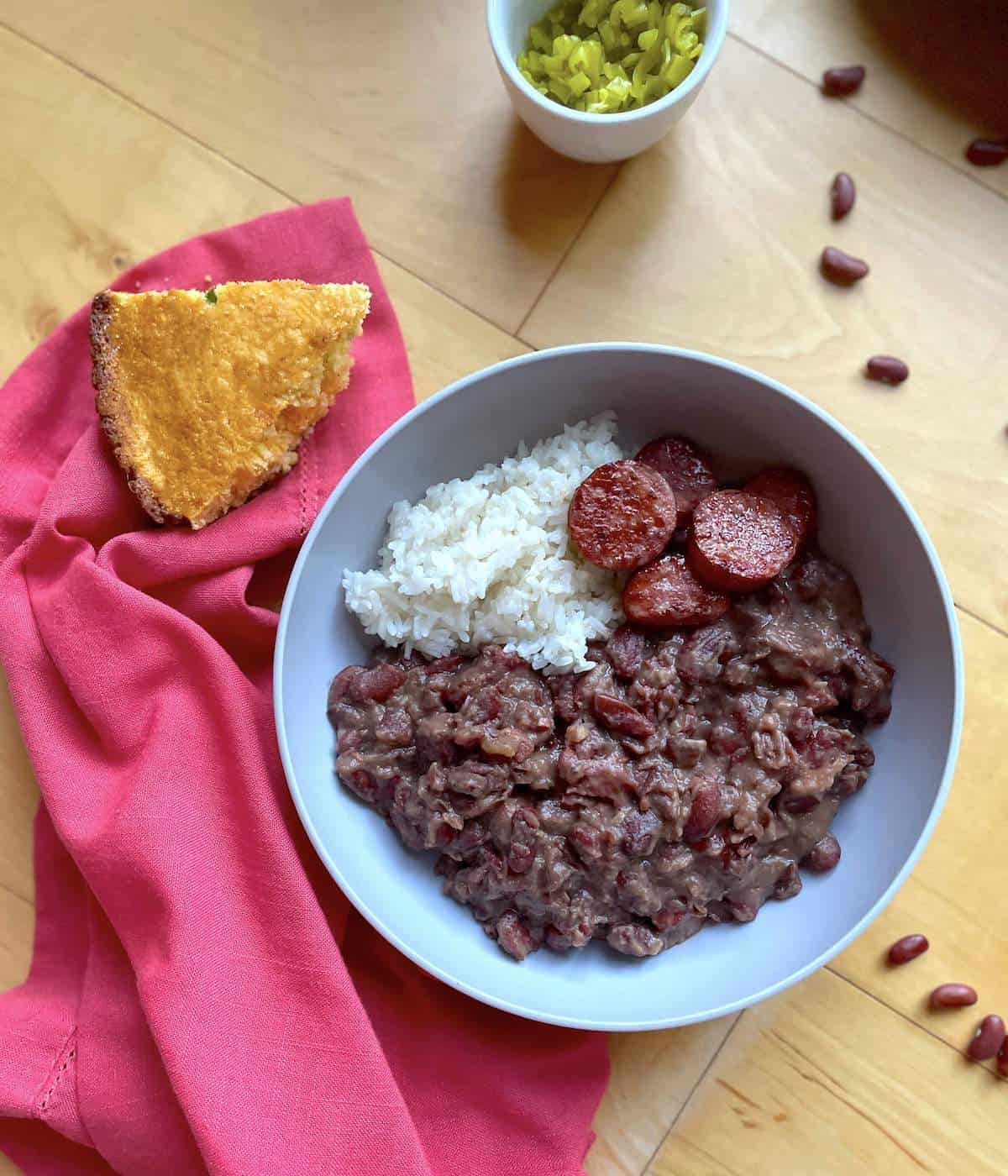 A bowl of red beans and rice with sliced andouille sausage, a slice of cornbread on the side, and a pink napkin on the side.