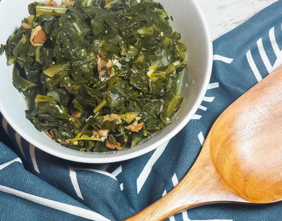A white bowl of collard greens with bits of bacon on a blue and white towel with a wooden spoon.