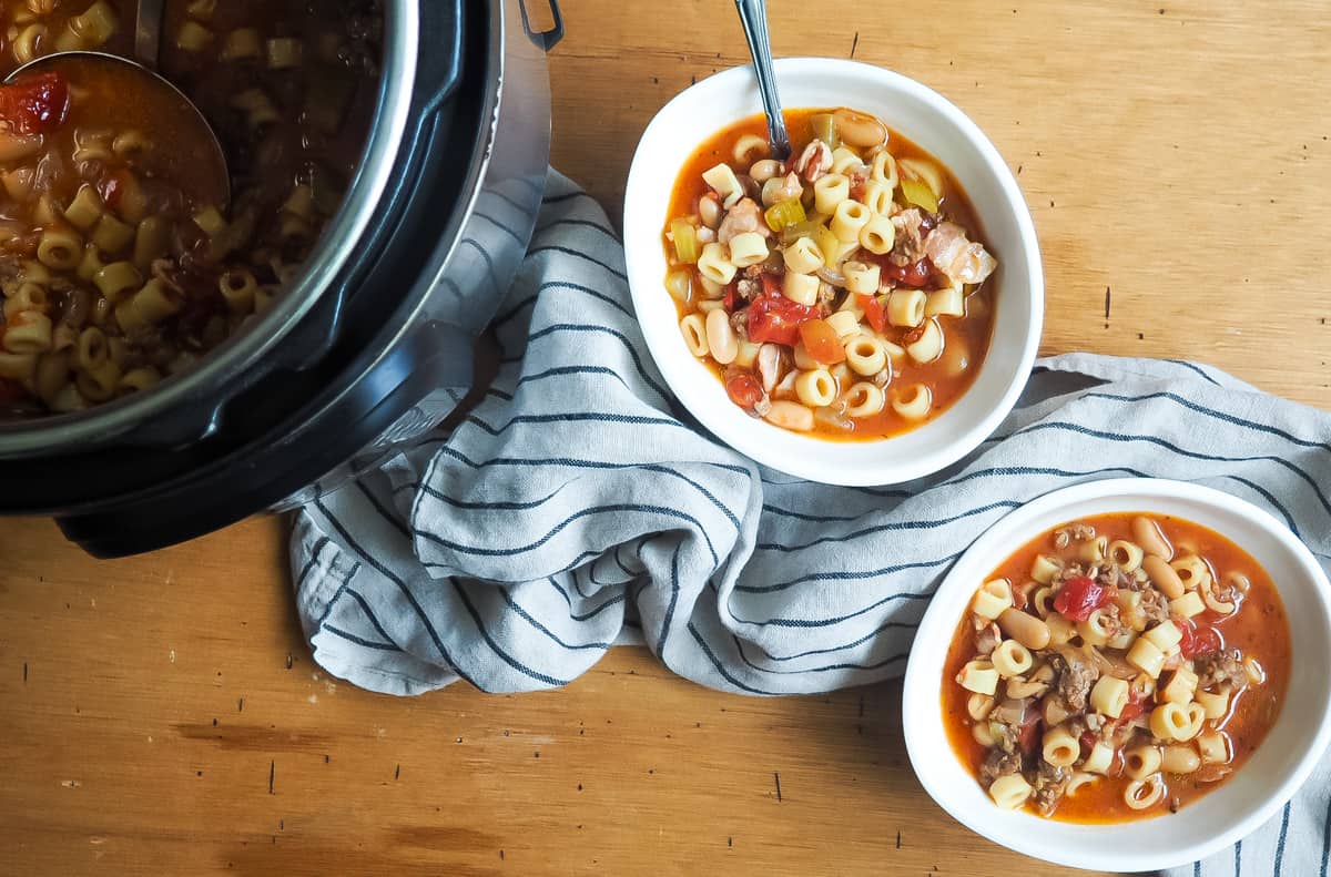 Two white bowls of pasta e fagioli next to an instant pot with a gray and blue dishtowel.