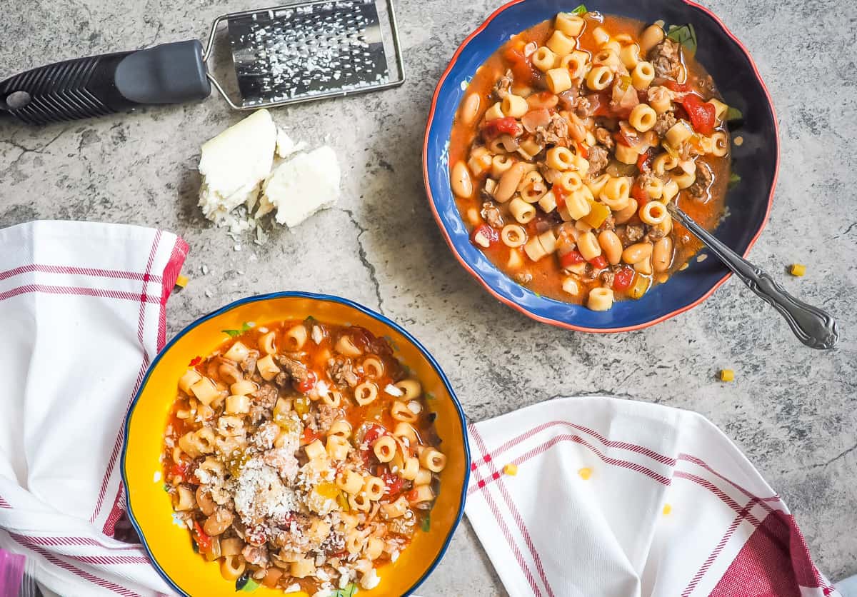 Yellow and blue bowls of pasta e fagioli with a spoon and a block of parmesan cheese.