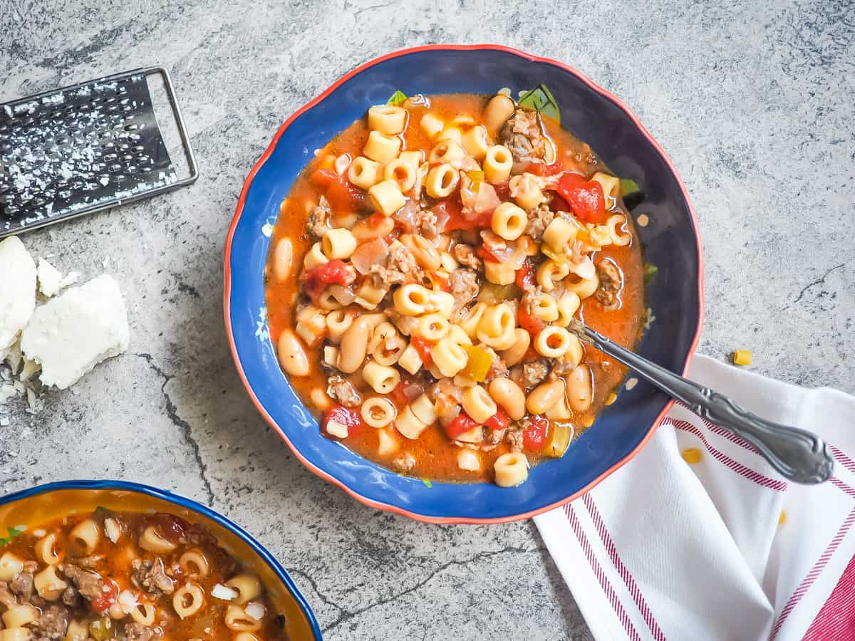 A blue bowl of pasta e fagioli with a spoon and a block of parmesan cheese.