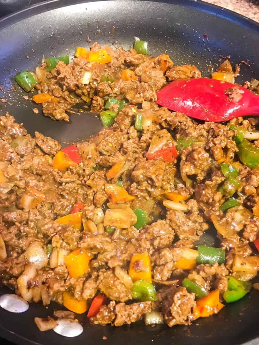 A frying pan with taco meat sauteing with peppers and onions and a red rubber spatula.