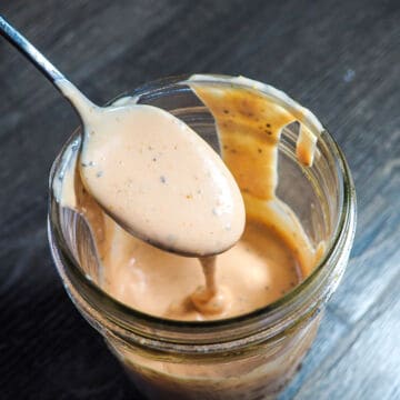 A jar of light orange Raising Cane's dipping sauce with a metal spoon on a gray wooden background.