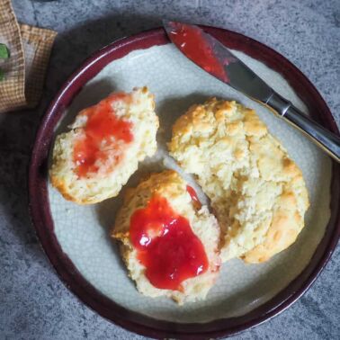 A drop biscuit cut in half with strawberry jam on top on a decorative plate.
