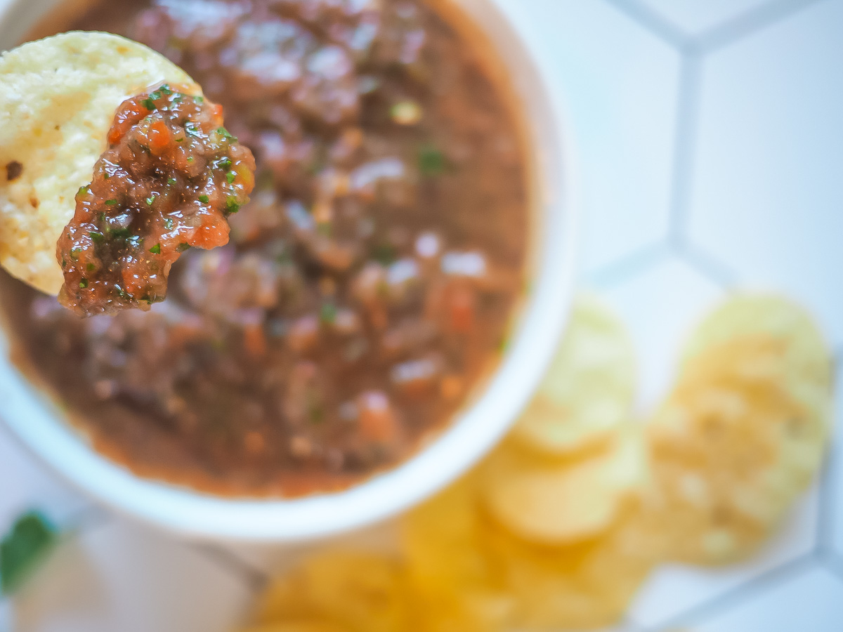 A chip with homemade salsa on it above a bowl of salsa and a pile of tortilla chips.
