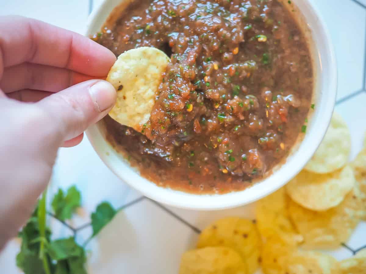 A hand dipping a chip with homemade salsa on it with a pile of chips and cilantro next to it.