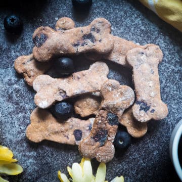 A pile of blueberry dog biscuits and blueberries on a gray background with a yellow flower and a yellow napkin.