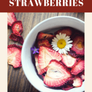 A pin image of bowl of dehydrated strawberries on a wood background with some daisies and small purple flowers scattered around.