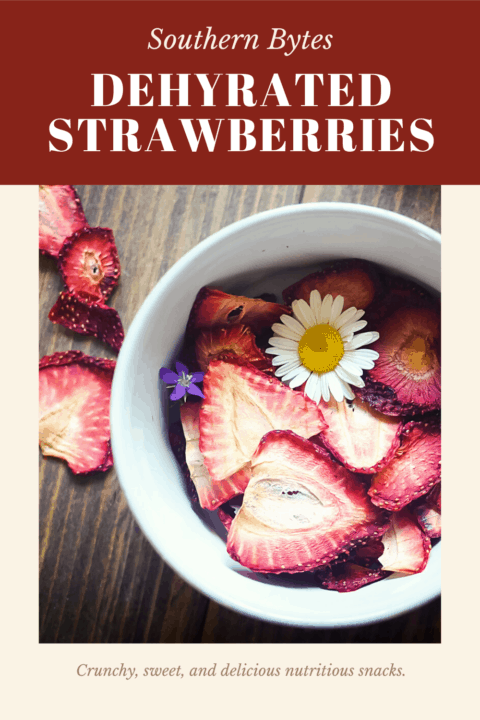 A pin image of bowl of dehydrated strawberries on a wood background with some daisies and small purple flowers scattered around.