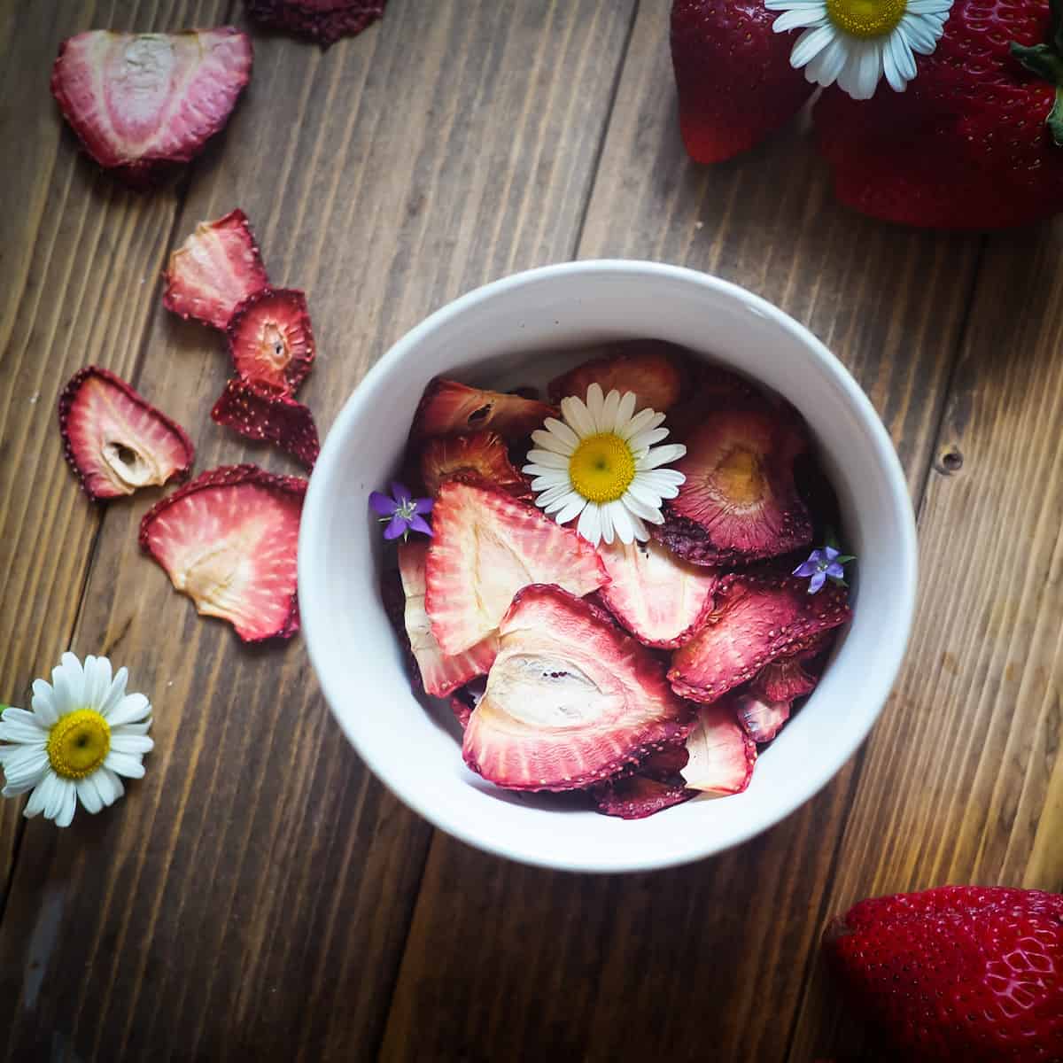 A bowl of dehydrated strawberries on a wood background with some daisies and small purple flowers scattered around.