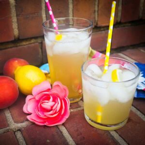 Two glasses of peach lemonade on brick steps with colored straws, peaches, a lemon, a pink flower, and lemon slices.