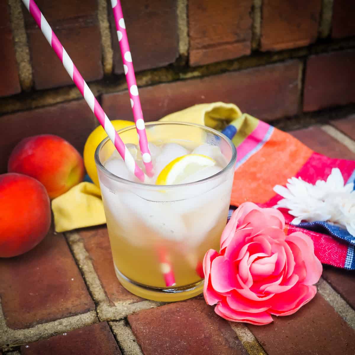 A glass of peach lemonade with two pink straws and a pink flower on a brick step.