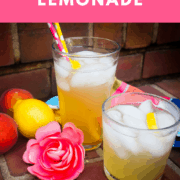 A pin image of two glasses of peach lemonade on brick steps with colored straws, peaches, a lemon, a pink flower, and lemon slices.