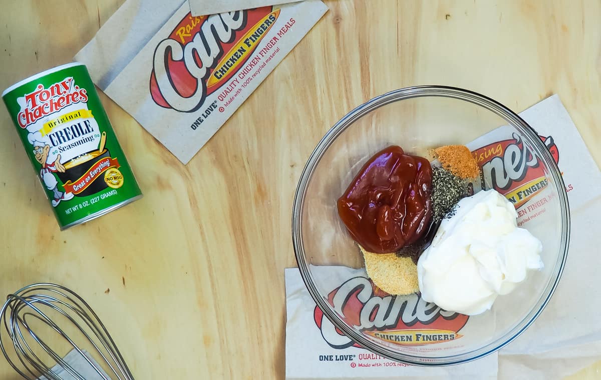 An unmixed bowl of Raising Cane's dipping sauce on a wood background with a whisk and container of Tony's Creole Seasoning next to it.