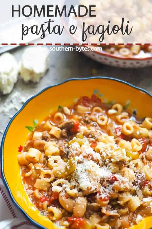 A pin image of a blue bowl of pasta e fagioli with a block of parmesan cheese on a gray background.