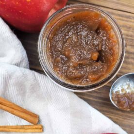 a jar of apple butter with a dish towel, a spoon, and an apple.