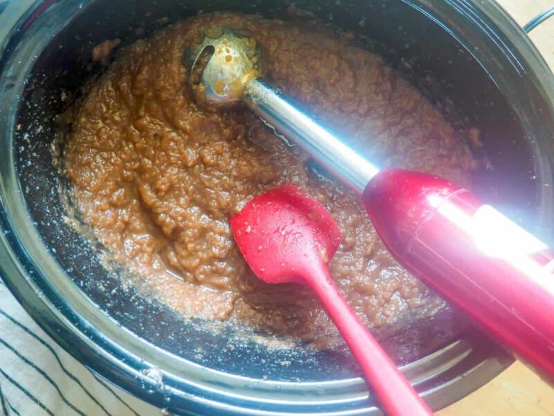 a crockpot with apple butter, an immersion blender, and a red spatula in it.