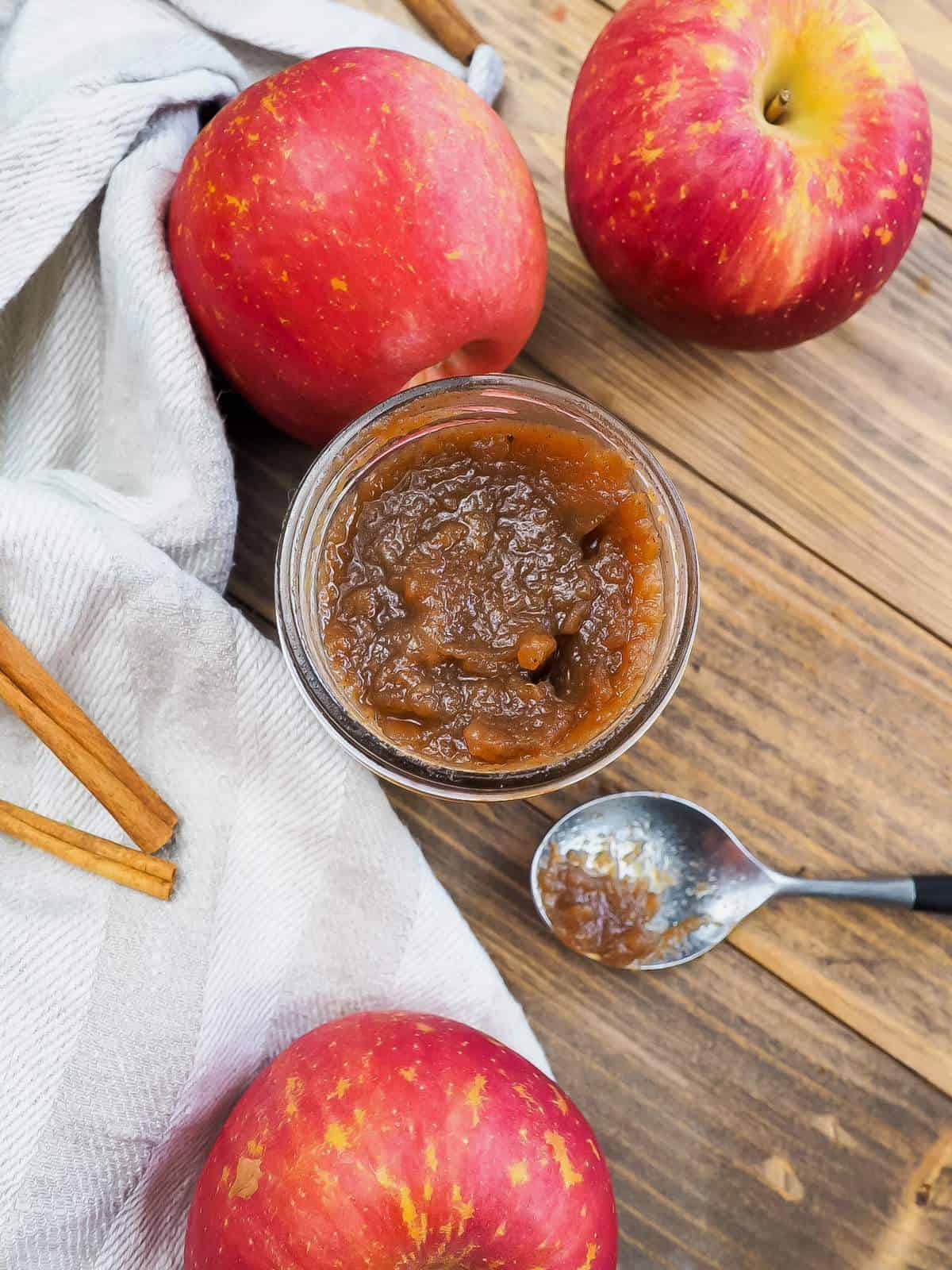 a jar of apple butter with a dish towel, a spoon, cinnamon sticks, and some apples.