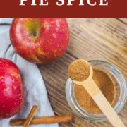 A pin image of a jar of apple pie spice with a small wooden spoon on top of the jar and some apples in the background.