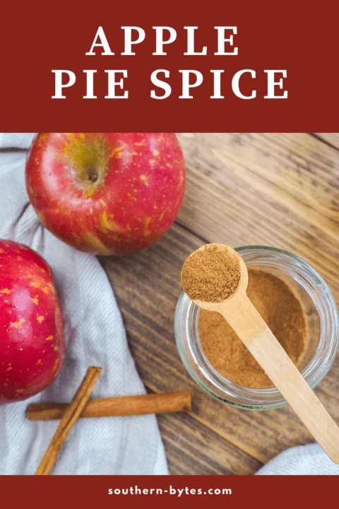 A pin image of a jar of apple pie spice with a small wooden spoon on top of the jar and some apples in the background.