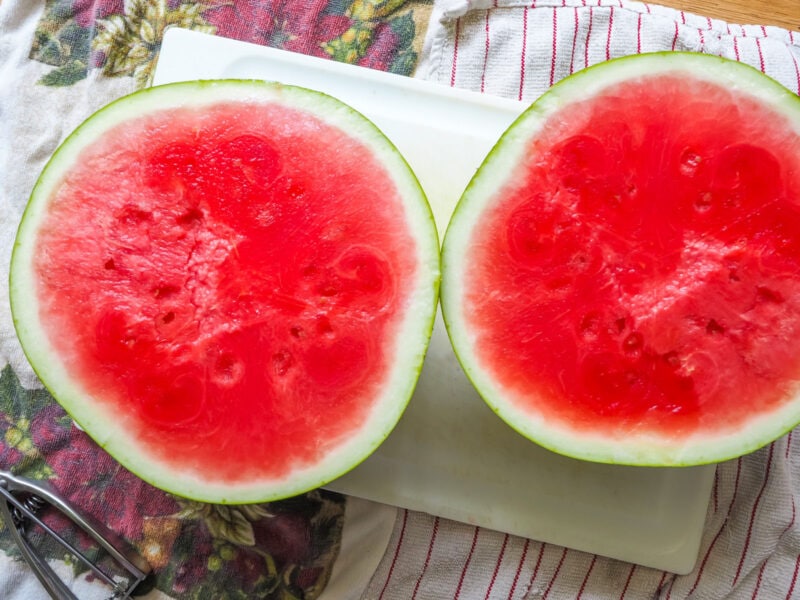 A watermelon cut in half on a white cutting board.