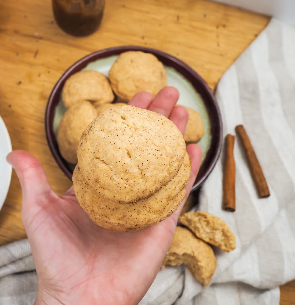 A hand holding a pile of snickerdoodles over a plate of cookies and two cinnamon sticks.
