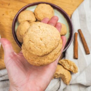 A hand holding two snickerdoodles over a plate of cookies and two cinnamon sticks.