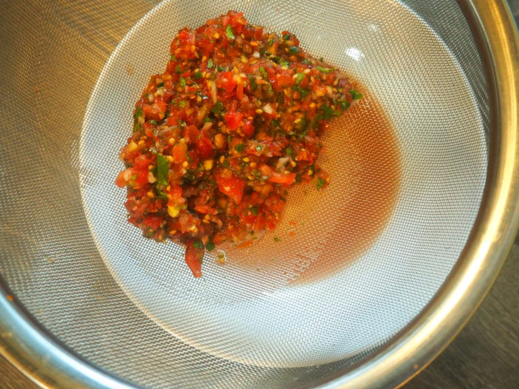 a metal strainer with salsa in it, draining liquid into a white bowl.