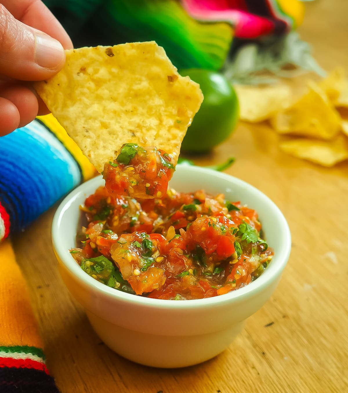 A chip dipping into salsa in a small white bowl and a lime and chips in the background.