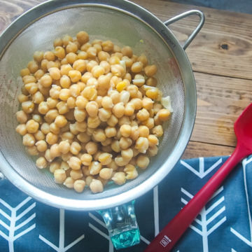 Cooked chickpeas in a metal strainer with a blue napkin and a red rubber spatula.