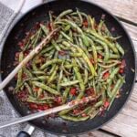 A frying pan of peruvian green beans with bell pepper and onions and a pair of tongs.