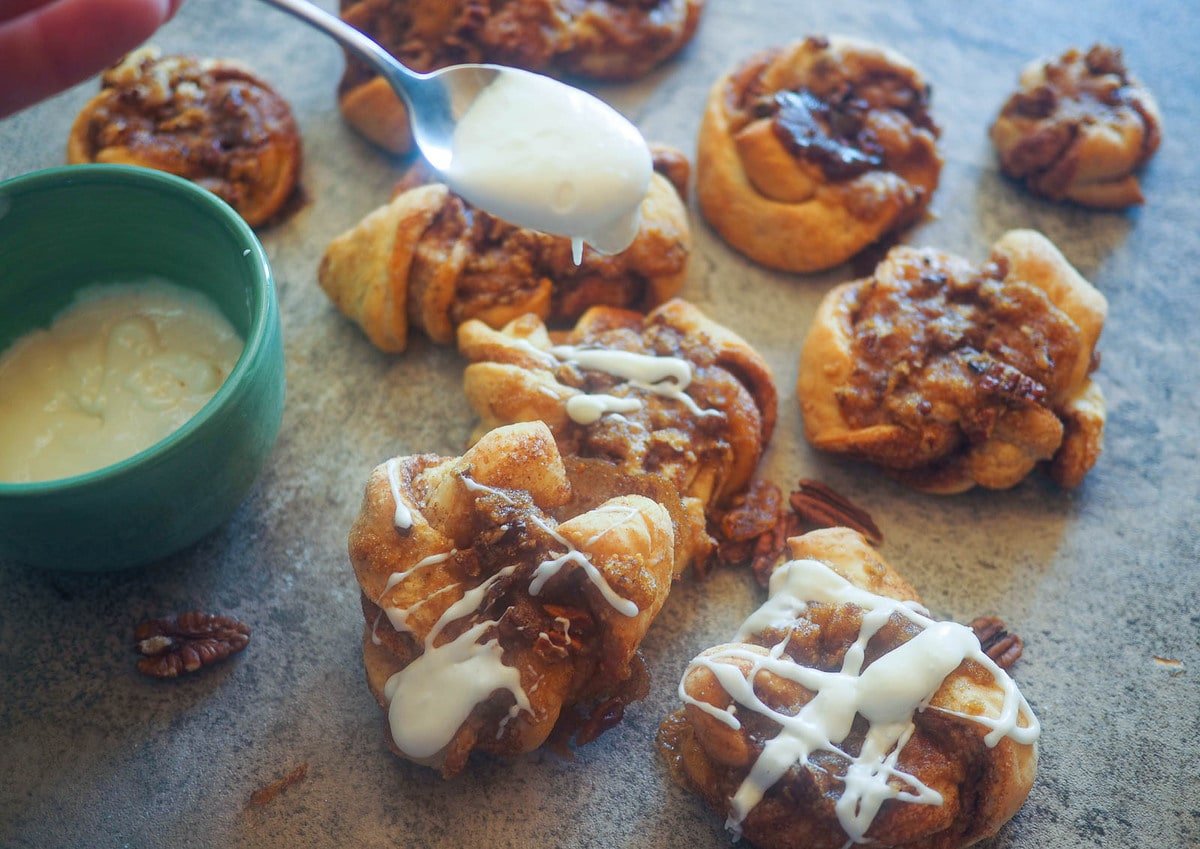 Praline crunch cinnamon knots on a gray board and a spoonful of icing being poured on them.