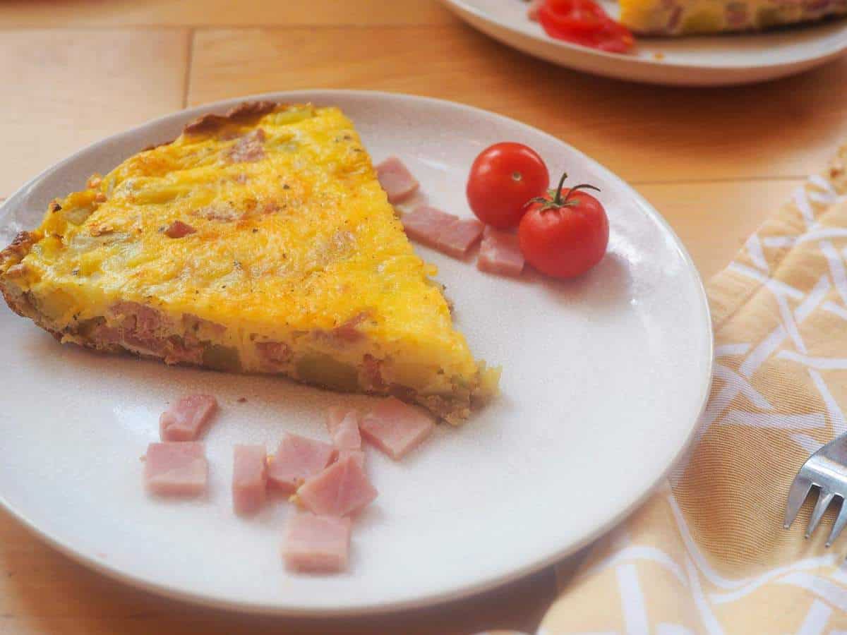 A slice of ham and gruyere frittata with some fresh tomatoes on a white plate.