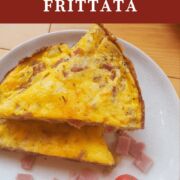 A pin image of two stacked slices of ham and gruyere frittata with some fresh tomatoes on a white plate.