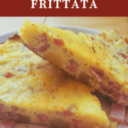 A pin image of two stacked slices of ham and gruyere frittata with some fresh tomatoes on a white plate.