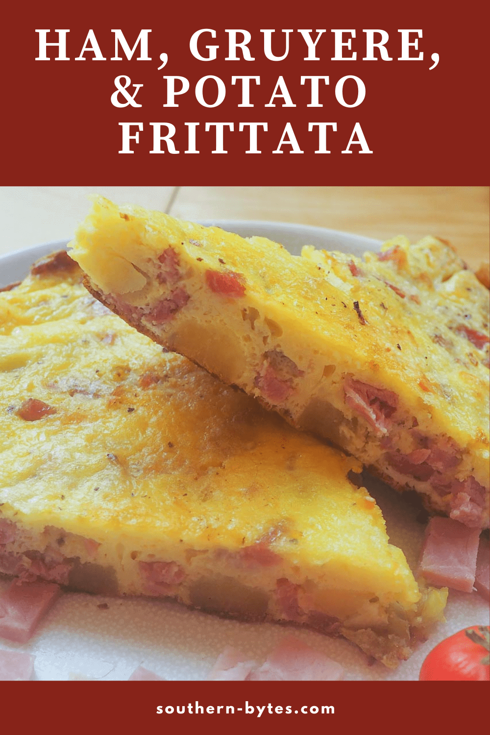 A pin image of two stacked slices of ham and gruyere frittata with some fresh tomatoes on a white plate.