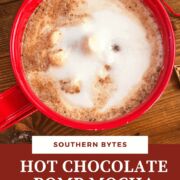 A pin image of a zoomed in red mug of hot chocolate topped with steamed milk on a wooden background surrounded by spilled coffee beans.