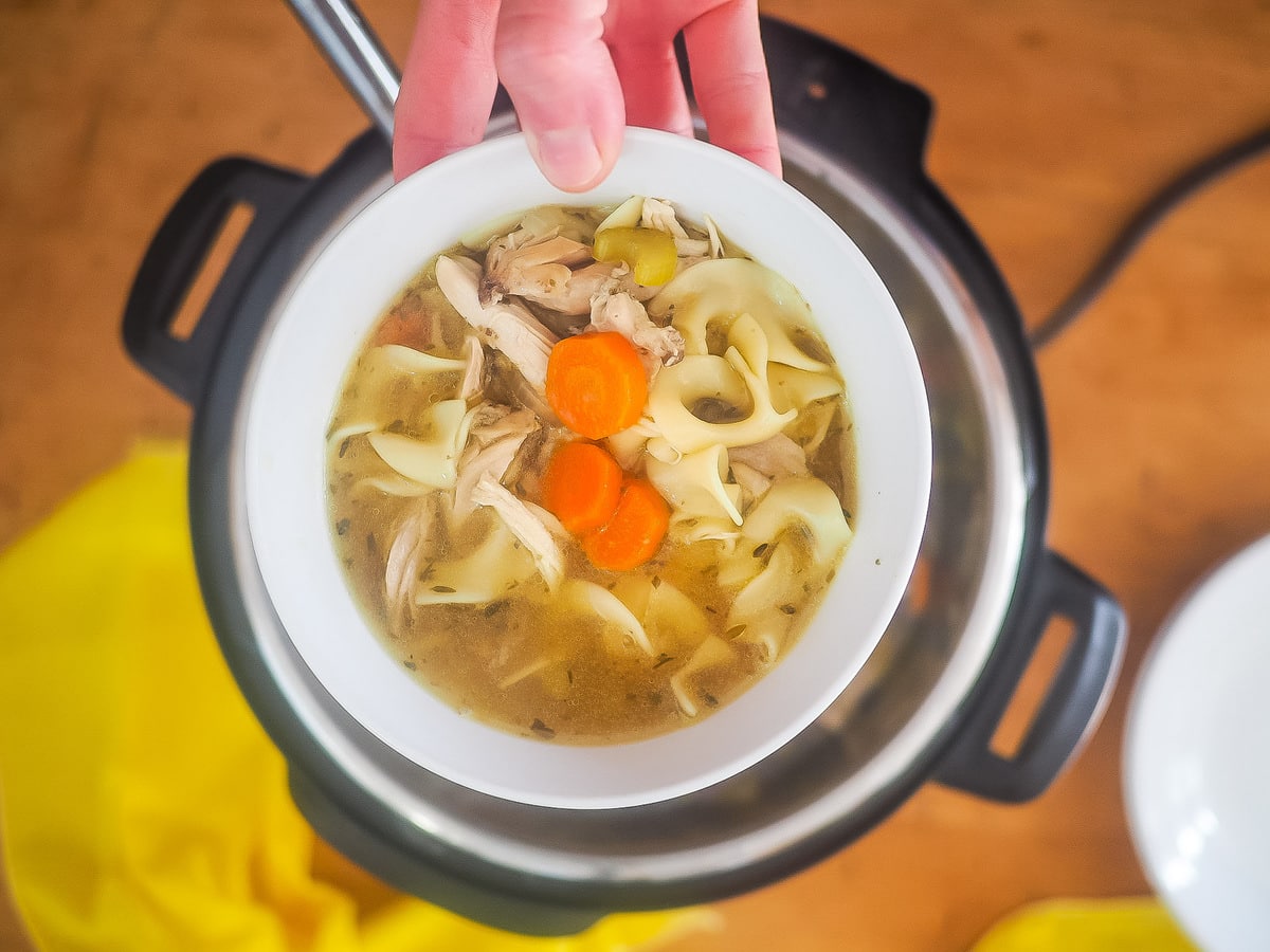 An hand holding a bowl of instant pot chicken noodle soup over an instant pot.