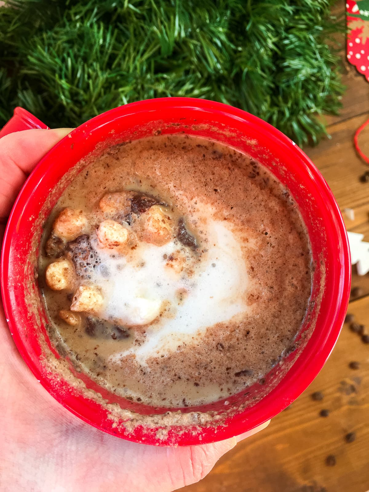A zoomed out image of a hand holding a red mug of hot cocoa with green garland in the background.