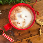 A red mug of hot chocolate topped with steamed milk on a wooden background surrounded by spilled coffee beans.