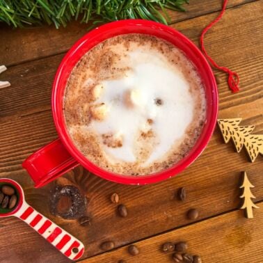 A red mug of hot chocolate topped with steamed milk on a wooden background surrounded by spilled coffee beans.