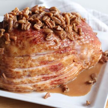 A sliced ham on a white plate with maple, bourbon, and pecan glaze drizzled over the top of it.