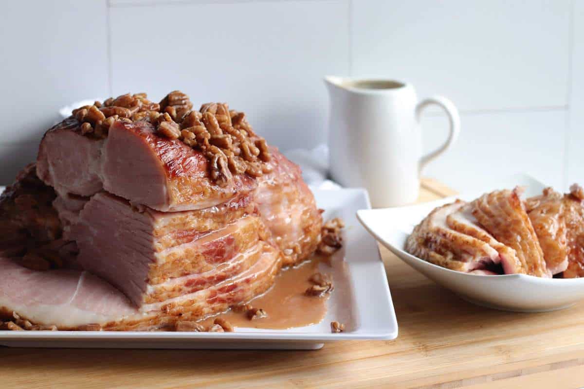 A sliced ham on a white plate with maple, bourbon, and pecan glaze drizzled over the top of it with slices off to the side.