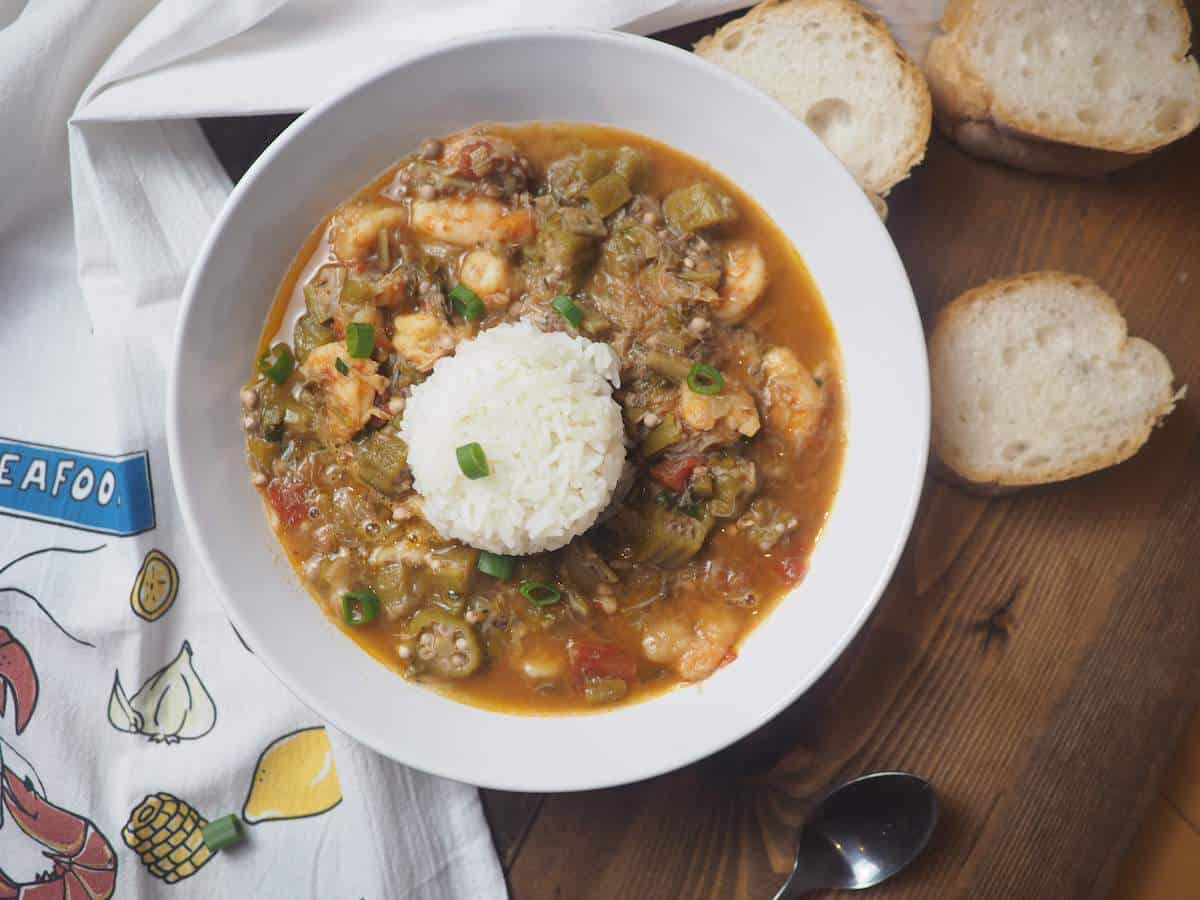 A white bowl of shrimp and crab seafood gumbo with rice, green onions, and french bread.
