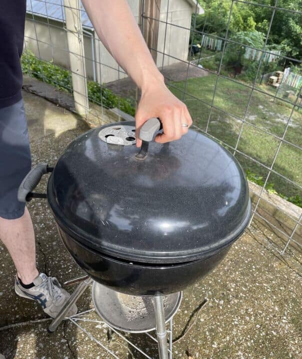 A charcoal grill with the lid on and a man holding onto the lid.