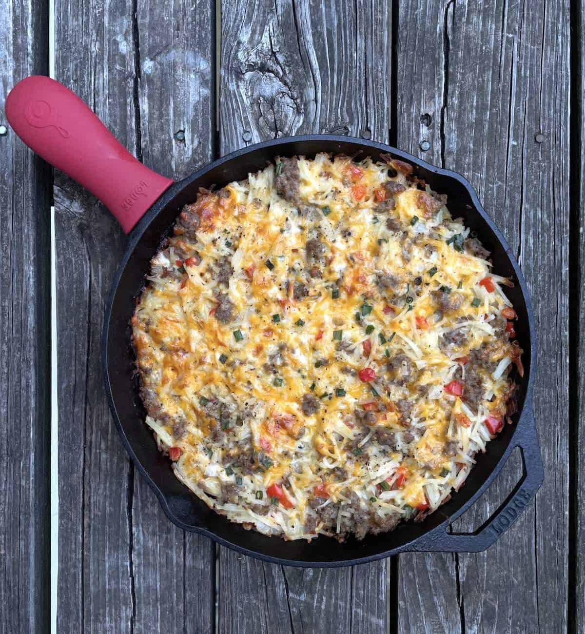A cast iron pan filled with cheesy hash brown casserole on a wood background.