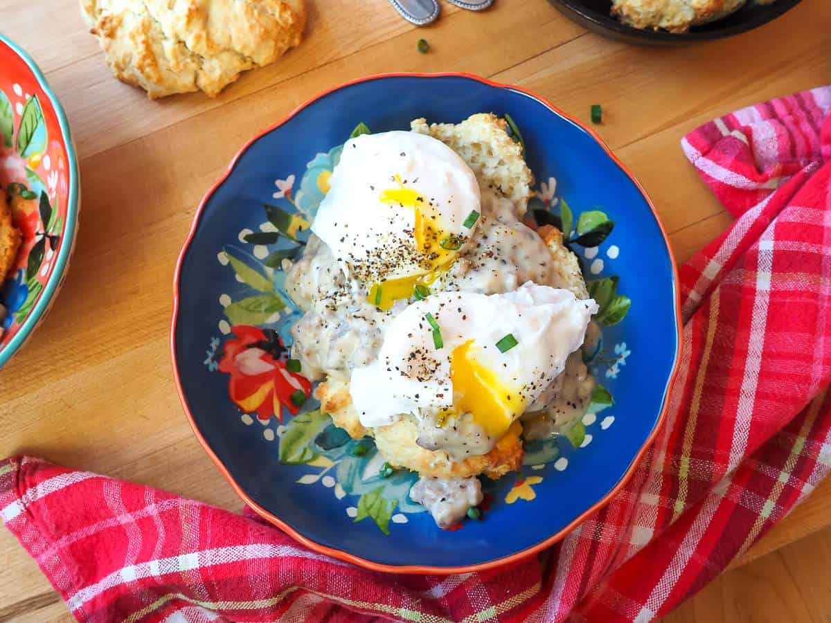 A blue bowl with two biscuits topped with sausage gravy and two poached eggs.