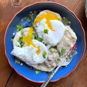 A blue bowl with two biscuits topped with sausage gravy and two poached eggs.
