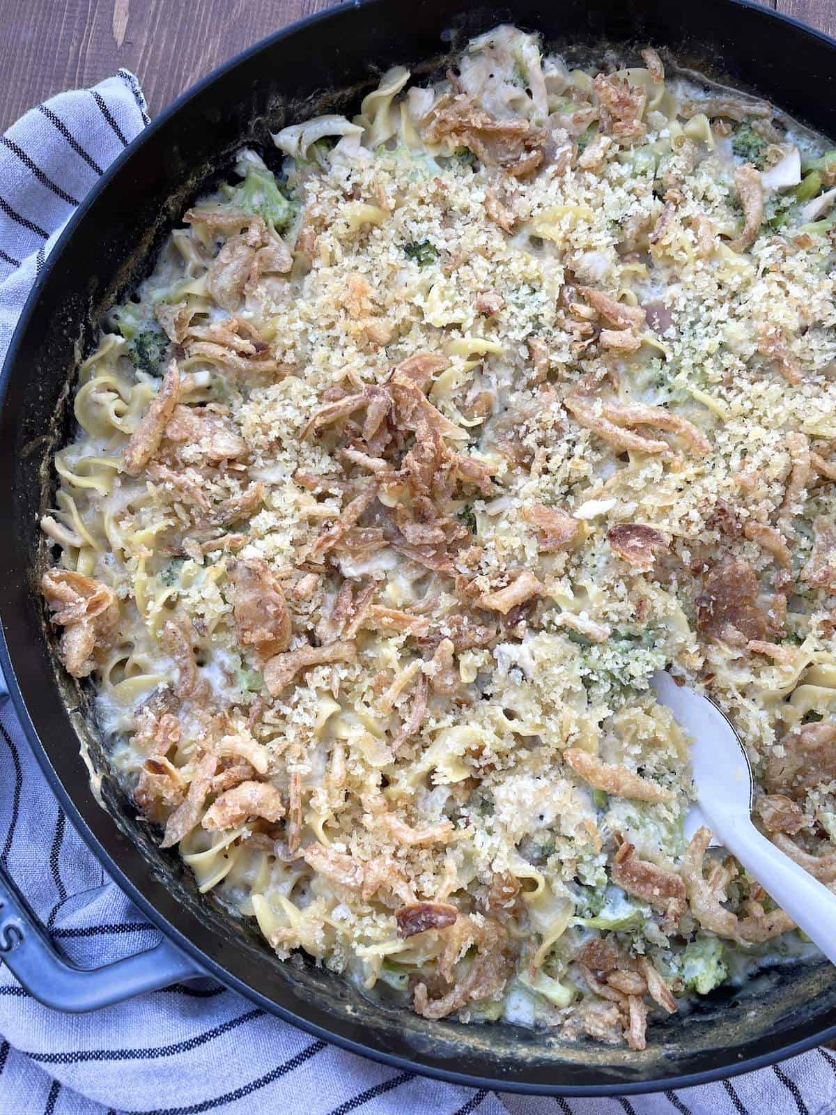A large cast iron pan filled with cheesy chicken and broccoli noodle casserole with a white serving spoon.