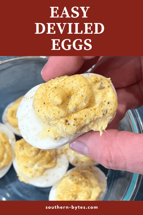 A pin image of a hand holding a deviled egg over a bowl of eggs.